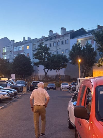 The image shows a street scene in Lisbon, Portugal, at dusk. A man with white hair, wearing a light-colored short-sleeved shirt and khaki pants, is walking away from the camera. He is on a paved street lined with parked cars. To the left of the street, a multi-story apartment building with many windows is visible. Some windows are illuminated, suggesting people are home. Trees are interspersed between the building and the street, with some branches reaching towards the building.
In the foreground, an orange car is parked, and further down the street, other cars are parked in a line, including a silver car and several darker-colored vehicles. The streetlights are on, casting a warm glow. The sky is a clear, pale blue, indicating the transition from day to night. The overall atmosphere is calm and residential.