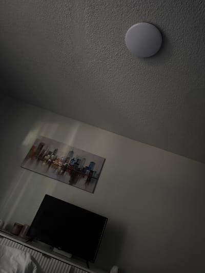 The image shows an interior view of a room, likely a bedroom or living area, with a textured ceiling that has a round, white ceiling light fixture. On the wall below the ceiling, there is a framed canvas print depicting a city skyline at night. To the right of the painting and lower on the wall, a black flat-screen television is visible, resting on a white cabinet or shelf. The room is dimly lit, with some light casting streaks on the wall from an unseen source, possibly a window. The overall impression is one of a modern, lived-in space. The image does not provide any specific clues about its location in North Highlands, United States.