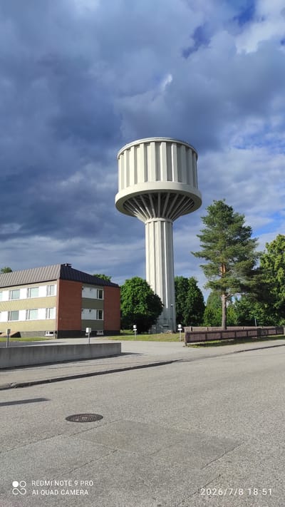 The image shows a water tower and a building against a cloudy sky. The water tower is tall and cylindrical with a distinctive ribbed base and a wider top section. A multi-story building with red brick and light-colored sections is visible next to the water tower. Trees are present in the background, and a wooden fence runs along the side of a paved road in the foreground. The time of day appears to be late afternoon or early evening, given the light and shadows, and the weather is partly cloudy with dramatic cloud formations.

Visible text on the image includes: "2026/7/8 18:51" and "REDMI NOTE 9 PRO AI QUAD CAMERA". The location is identified as Iisalmi, Finland. There are no people or specific activities depicted in the image. The scene is an outdoor view of urban infrastructure and natural elements under a dynamic sky.