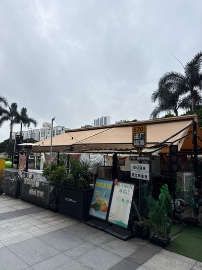The image shows a street-level view of an outdoor dining or drinking establishment, likely a bar or cafe, under a light brown awning. The establishment is situated in Haizhu District, China. In the foreground, there is a tiled sidewalk and several planters with various green plants, including palm-like foliage. Several signs are visible. One prominent yellow and black sign reads "520 进唱 喝酒" with smaller text below. Another sign below this states "广州·琶洲保利站". Other signs show text in Chinese characters and English, advertising "COCKTA" and "很大杯BIG 冰美式" with a price of "¥12.9". To the left, a wire mesh structure displays a sign with "YOJO mini 琶洲". In the background, several tall apartment buildings and palm trees are visible against an overcast sky. There are no people directly visible in the frame. The scene appears to be an urban commercial area.