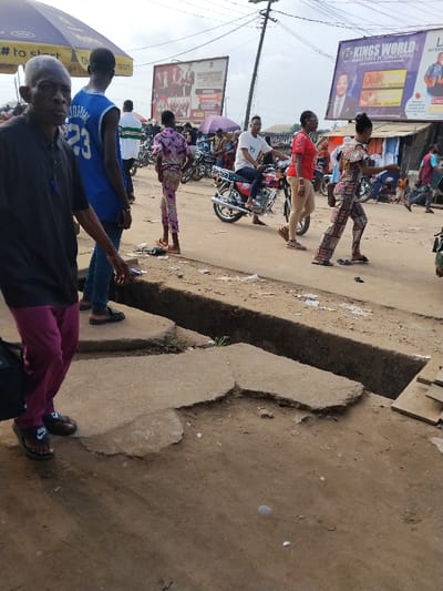 The image depicts a busy street scene in Umuode, Nigeria. In the foreground, an open concrete drainage ditch runs horizontally across the frame, flanked by broken concrete slabs and reddish-brown soil. On the left, an older man with short gray hair, wearing a black polo shirt, maroon trousers, and black Nike slide sandals, holds a black shoulder bag and looks towards the viewer. Behind him, a younger man in a blue sleeveless basketball jersey with "23" and "JORDAN" on the back, dark blue jeans, and brown shoes faces away, looking towards the right side of the street.
Further into the scene, a person in a purple and pink patterned two-piece outfit stands barefoot on the unpaved roadside. Adjacent, a man in a white t-shirt and dark cap rides a red motorcycle, moving from right to left. A woman in a red t-shirt, khaki-colored trousers, and brown sandals walks alongside the motorcycle, also moving from right to left. To her right, another woman in a multicolored patterned jumpsuit walks with her arms slightly outstretched.
The background reveals a bustling street with several other individuals, some walking, some standing, and additional motorbikes parked or in motion. Prominent overhead are two large billboards: one on the left for "BUCKINGHAM COLLEGE HILLTOP CITY SCHOOL" displaying multiple students, and another on the right for "KINGS WORLD MINISTRIES INTERNATIONAL" featuring a man in a suit and listing service times. A utility pole with overhead power lines stands near the center-right. The sky is overcast.