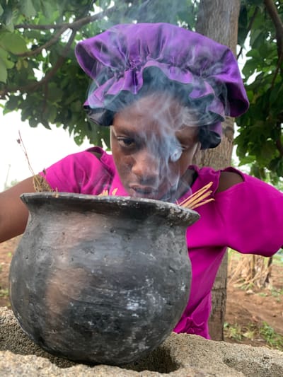 In Agban, Nigeria, a woman wearing a magenta top and purple headscarf is positioned outdoors. She is situated next to a rough concrete structure. In front of her, a black, spherical pot emits smoke; thin sticks are placed near the pot's opening. Trees with green foliage are visible in the background.