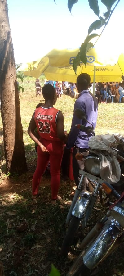 An outdoor gathering is depicted in Burundu, Kenya, under bright, sunny daylight. In the foreground, a woman wearing a red sleeveless jersey with "LAKERS 23" on the back and red leggings stands with her back mostly to the viewer. Beside her, a man in a blue long-sleeved shirt also faces away. They stand near a large tree trunk on the left and motorcycle parts, including a headlight and chrome fender, in the lower right.
In the background, under a large yellow tent, numerous people are seated on blue and white plastic chairs, suggesting a social event or community gathering. The tent features visible branding and text, including "SMs WIN", "SMs", and ".com". The ground is grassy with patches of dirt and fallen leaves, indicating a natural, possibly rural or semi-rural setting. The strong sunlight casts clear shadows, suggesting it is daytime. The overall atmosphere appears casual and relaxed.