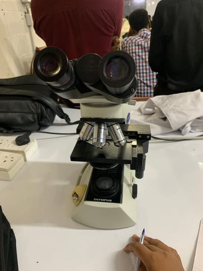 The image depicts an indoor laboratory or classroom setting in Dar es-Salaam, Tanzania. A beige and black Olympus binocular microscope, likely a CX series model, is centrally positioned on a white, reflective table. The microscope features two black eyepieces, a mechanical stage, and a revolving nosepiece displaying four objective lenses, one clearly marked "OLYMPUS 40x/0.65" and another "MPU." A black power cord extends from the microscope's base. To the left of the microscope, a black bag, possibly a shoulder bag or backpack, and two off-white electrical multi-sockets are visible on the table. In the bottom right foreground, a hand, belonging to an individual, holds a clear blue ballpoint pen over a white sheet of paper. The blurred background reveals parts of three individuals: one in a maroon garment, another in a blue and white checkered shirt, and a third in a black garment. A crumpled white lab coat or similar fabric lies on the table behind the microscope, and a tiled wall with white square tiles forms the furthest background. The scene suggests a practical laboratory or educational session in progress.