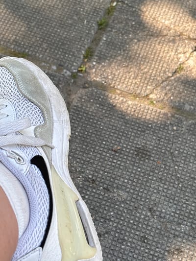 An overhead shot captures a person's foot adorned in a white sneaker, resting on a textured concrete pavement under bright daylight in Gotse Delchev, Bulgaria.
The primary subject is the right foot, partially visible, clad in a white athletic shoe. The shoe features a mesh upper, white laces, and a slightly worn-looking light gray suede-like panel around the toe and midfoot. A small black Nike swoosh logo is discernible on the lateral side of the shoe. The heel area incorporates a prominent translucent, yellowish-tinted air unit, typical of certain sneaker designs. Faint, stylized text, likely "AIR" or a branding element, is subtly molded into this unit. The white sole and upper show signs of everyday wear, with minor scuffs and a slightly dingy appearance in areas. A white sock is visible around the ankle, and a sliver of skin peeks out from the top of the shoe.
The setting is an outdoor paved surface, likely a sidewalk or path, composed of concrete pavers with a distinct grid or square pattern. Sections of the pavement are bathed in bright sunlight, while others are cast in shadow, suggesting an overhead obstruction like a tree or building. Small cracks in the concrete and sporadic patches of green grass or weeds add to the outdoor environment. The strong contrast between light and shadow indicates a sunny day, likely during morning or afternoon hours. No specific activity beyond standing or being present is depicted. There are no discernible emotions or clear textual elements beyond the faint branding on the shoe.