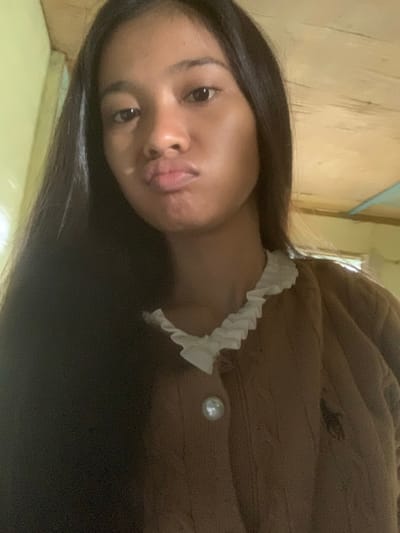 A young woman with dark hair is looking at the camera with her lips pursed. She is wearing a brown sweater with a white ruffled collar and a pearl button. The background appears to be indoors with a wooden ceiling. There are no visible landmarks or contextual clues to determine the specific location within Sungai Meriam, Indonesia.