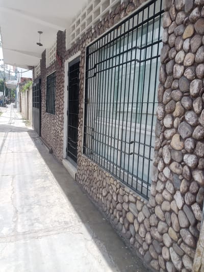 This image depicts the exterior of a building with a unique facade constructed from various sizes of rounded stones, giving it a rustic appearance. A sidewalk runs alongside the building, leading into a street scene in the background. The street appears to be in a residential or commercial area, with other buildings and some greenery visible.
In the foreground, the sidewalk is made of concrete slabs with visible cracks, indicating age or wear. The building's wall is prominently covered in the natural stones, interspersed with sections of white, lattice-like brickwork near the roofline. Black metal bars protect the windows, adding a sense of security. A simple black metal gate is visible on the left side of the building, suggesting an entrance. Overhead, a white awning extends from the building, and a single light fixture hangs from the ceiling of the awning.
The background shows a narrow street with power lines overhead. There are some vehicles and people faintly visible, suggesting some activity on the street. The overall lighting suggests it is daytime, likely with bright sunlight given the strong shadows cast on the sidewalk and street. The weather appears to be clear and fair. The scene gives a sense of an ordinary day in a South American town, possibly in Santa Ana, Venezuela, given the location context. No explicit text is visible in the image.