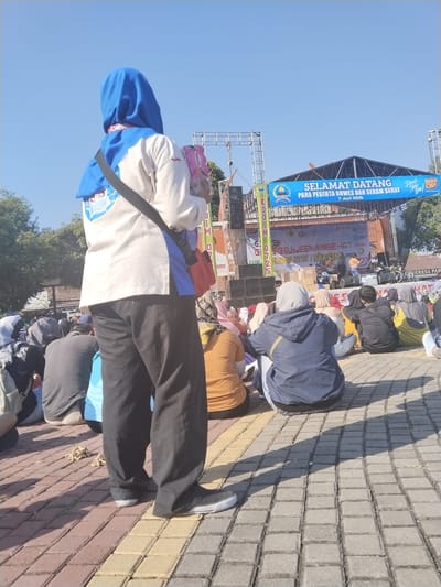 The image depicts an outdoor event taking place in Sragen, Indonesia, specifically referenced as "KODIM 0725 / SRAGEN" on a stage banner. The scene is observed from a low angle, with a prominent individual in the foreground, viewed from the back. This individual wears a blue hijab, a white long-sleeved shirt with blue accents, dark pants, and black sneakers with white soles. A black shoulder strap crosses their back, and they hold a pink multi-pack of Yakult.
The foreground person stands on a paved surface composed of reddish-brown and gray interlocking pavers, which extends into the middle ground where numerous other individuals are seated directly on the ground, facing a stage. These attendees are primarily seen from behind, many wearing hijabs or hooded garments.
In the background, a raised stage is visible, featuring a white backdrop banner displaying logos (including Fastlink and BRI) and text identifying the event as "Gowes & Senam Sehat" (Cycling & Healthy Exercise) by "KODIM 0725 / SRAGEN." Performers or presenters are discernible on the stage, with musical instruments such as drums present. To the left and right of the stage, metal truss structures support sound equipment, lights, and a large blue overhead banner that reads "SELAMAT DATANG PARA PESERTA GOWES DAN SENAM SEHAT 7 Juni 2026," along with "Hydro Coco" and "Don't Stop Now!" branding. Trees are visible on the left and right edges of the background. The sky is clear and blue, indicating a sunny day, with strong shadows cast by the people and structures on the pavement.
