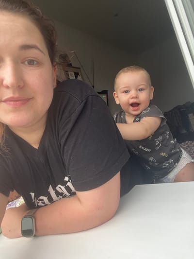 The image shows a woman and a baby in an indoor setting. The woman is in the foreground, with her face partially visible, looking towards the camera. She is wearing a black t-shirt and a watch on her left wrist. The baby is behind her, leaning forward and looking at the camera with a wide smile. The baby is wearing a grey onesie with a pattern of cartoon characters and the words "ENJOY!" and "PLAY!", and a diaper. They appear to be sitting at a white surface, possibly a table, with a window or opening in the background. The environment is dimly lit, suggesting it is indoors. There are no specific indicators in the image that would allow for the determination of its location in Chelyabinsk, Russia.