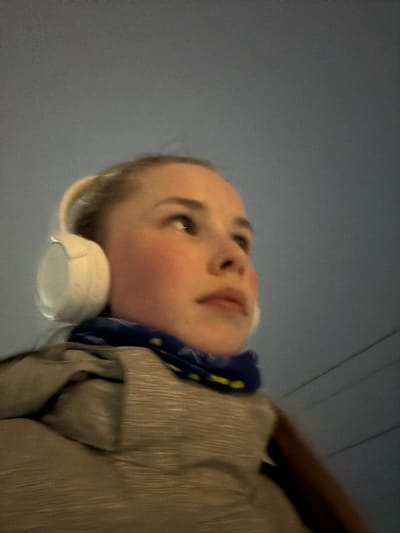 A close-up, slightly blurred, upward-angled shot depicts a person with fair skin and blondish hair wearing white headphones. The person is also wearing a blue scarf with yellow accents around their neck and a tan jacket or coat. The background is a blurred, light blue-grey, possibly indicating the sky or an indoor ceiling. The location cannot be determined definitively as Berezniki, Russia, without additional contextual information. No specific actions or interactions are occurring within the visible frame. The environment is indeterminate, suggested only by the background color.