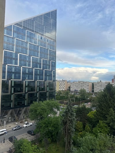 The image shows a view of Warsaw, Poland, focusing on a modern, angular glass building in the foreground. The building has a unique geometric design with many windows reflecting the cloudy sky. To the right of the modern building, there are several older, low-rise apartment buildings typical of Soviet-era architecture. A dense area of trees and greenery separates the modern building from the older apartments. A street with several cars is visible in front of the modern building, and a railway track with overhead wires runs through the trees further in the background. The sky is partly cloudy with a mix of blue sky and large, white clouds, suggesting it might be a slightly overcast day.