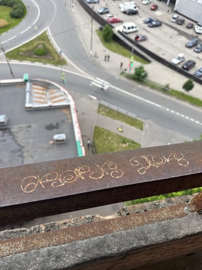 The image shows a high-angle view of a street scene in Korolyov, Russia, with a rusty metal railing in the foreground. The railing has graffiti on it, which appears to be in Cyrillic letters and is gold in color. The background shows a road with several cars, a roundabout with green landscaping, and some buildings. There are also some pedestrians visible on the sidewalk and crossing the street. The overall impression is that of an urban environment.