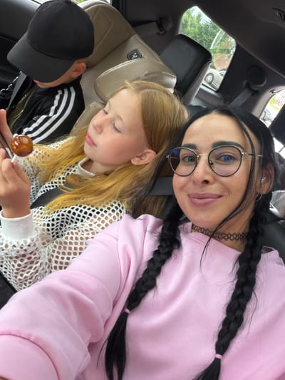 In the passenger seat of a vehicle, a young boy wearing a black baseball cap and a black and white striped jacket is visible, his face partially obscured by the cap. Beside him, a young girl with blonde hair and closed eyes holds a lollipop in her right hand. In the foreground, a woman with dark hair styled in two braids, wearing glasses and a pink sweatshirt, smiles at the camera. The interior of the car suggests a casual outing, possibly during a trip within the city of Veliky Novgorod, Russia.