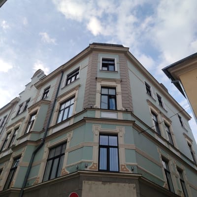 The image shows a pale teal-colored building with ornate light beige trim, viewed from a low angle looking upwards. The building has multiple floors and numerous dark-framed windows. The architectural style features decorative elements around the windows, horizontal banding, and angled projections or cornices. A downspout runs vertically along one side of the building. The sky above is a mix of pale blue with scattered white clouds. To the right, a sliver of a yellow building is visible. The scene suggests an urban environment, likely a historic district given the architectural style. The image was taken in Riga, Latvia.