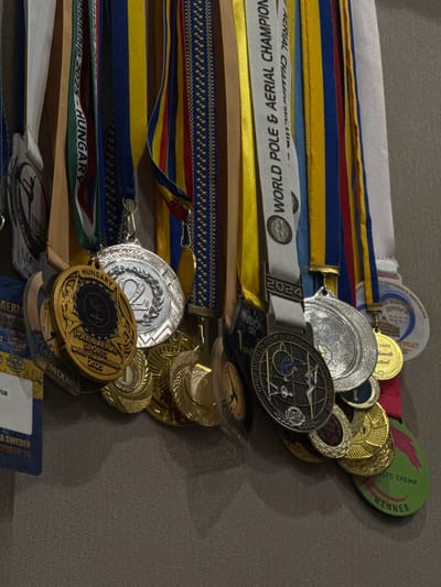 A close-up, head-on shot shows a collection of medals hanging against a textured, gray background, likely a wall. The medals, various sizes and colors, are suspended by ribbons of different patterns and hues, including blue, yellow, red, and white. Some ribbons bear text, such as "WORLD POLE & AERIAL CHAMPIONSHIPS" and "HUNGARY." Several medals have inscriptions and graphics, including numbers like "2" and "2025." One medal features a depiction of pole dancers and the text "POLE & AERIAL SPORTS WORLD CHAMPIONSHIP 2025." Another medal is green and oval-shaped with pink text that reads "WINNER." There's a partially visible blue tag with white text that says "SHOWDOWN SWEDEN OCTOBER 24". The overall impression is of an award display, possibly in a competition venue or a personal collection. The specific location in the city of Dnipro, Ukraine, is not evident from the image itself.