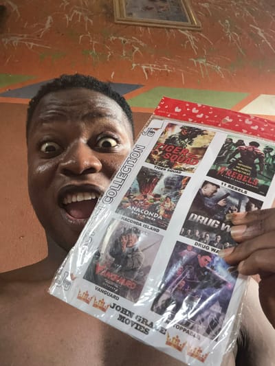 A close-up, rotated shot shows a man holding a movie collection package. The collection includes titles like "11 Rebels," "Drug War," "Tiger Squad," "Anaconda Island," and "Vanguard" featuring Jackie Chan. The collection is labeled "John Grace Movies." The package has "Made in China" printed on it and a small sticker with "JP" on the bottom corner. The man's face is partially obscured by the movie package, revealing his open mouth and teeth. The background features a wall painted with orange and green patterns and a framed picture.
