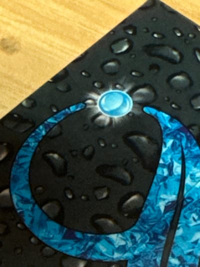 The image features a dark fabric surface, possibly a piece of clothing or a textile, with a pattern of simulated water droplets scattered across it. A prominent, stylized 'c' shape in shades of blue with a fractured ice-like pattern is visible. A single, larger, luminous blue sphere with radiating light streaks is positioned close to the 'c' shape. The background is a light tan surface, possibly a table or countertop. No people or specific objects indicating a location in Jiulongpo District, China, are discernible.