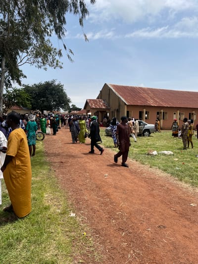 The image depicts a gathering of people on a dirt path in Durumi, Nigeria. A long, light brown building with a rusty, corrugated metal roof and open windows dominates the background on the right. Several individuals, dressed in vibrant, traditional attire, are walking or standing on the path and surrounding grassy area. Some are in groups, suggesting conversation or social interaction. A silver car is parked near the building. The environment is outdoors, with trees and a cloudy sky visible. The scene appears to be a community event or gathering.