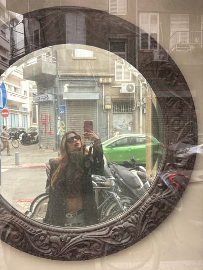 A young woman is depicted in a selfie, reflected in an ornate, dark-framed oval mirror, likely part of a storefront display. She stands facing the mirror, holding a light-colored smartphone aloft with her right hand to capture the image. Her left arm hangs casually, holding a dark strap. She wears dark sunglasses, a sheer dark top (possibly leopard print) exposing her midriff, and light-colored jeans with a distinctive belt buckle. Her long, light brown hair cascades over her shoulders. Her lips are slightly pursed in a pose.
The mirror's reflection reveals an urban street scene in Tel Aviv-Yafo, Israel, specifically identifiable by Hebrew signage including "סופר קאמרה" (Super Camera) and "ת.ד." (T.D. - likely an address marker), consistent with Allenby Street. The environment features multi-story, aged buildings with balconies, visible air conditioning units, and a mix of open and shuttered windows. One building's ground floor features roll-down metal shutters covered in graffiti. On the street, vehicles include a bright green hatchback car, multiple parked motorcycles, and bicycles. A man is visible walking on the sidewalk in the background, near a parked bicycle. Street furniture includes a blue circular sign with a white left-pointing arrow and a red circular "no entry" sign. The overall scene is captured in daylight, presenting a bustling urban streetscape with an overlay of a faint reflection of a building above the mirror's frame.