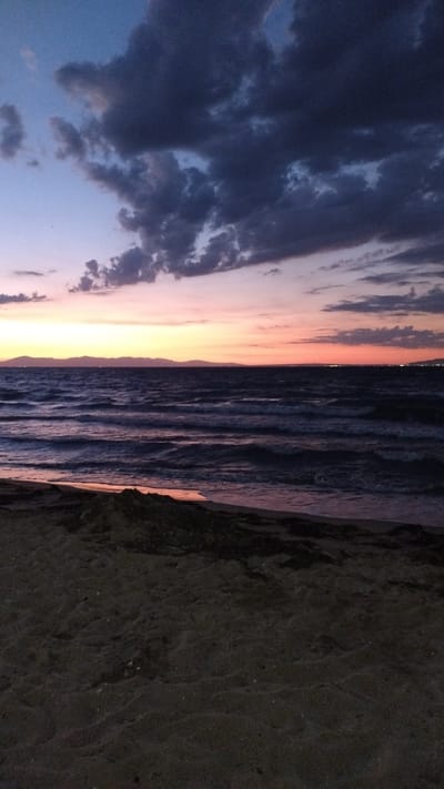 The image depicts a sunset over the Aegean Sea in Perea, Greece. The foreground is dominated by a dark, sandy beach with some scattered debris. The sea stretches out into the mid-ground, with gentle waves reflecting the colors of the sky. In the distance, a range of mountains forms the horizon, with faint lights suggesting a distant settlement. The sky transitions from a deep blue at the top to vibrant hues of orange and pink near the horizon, punctuated by dramatic, dark clouds. No people or animals are visible in the scene.