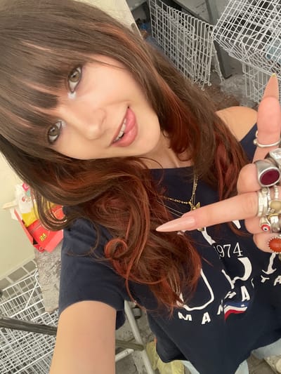 A young woman with brown hair with red undertones and bangs takes a selfie in what appears to be a storage area. She is wearing a dark blue t-shirt with white lettering that includes "1974" and "MAN". Her right hand is raised, showing several rings on her fingers and a long, pointed, light-colored fingernail. The background is cluttered with white wire shelves, some of which appear to be storage cages or shopping carts, and a red box with yellow labeling that says "24 PACKS". The lighting is somewhat dim, suggesting an indoor, possibly industrial or commercial, environment. The image was taken in Raanana, Israel.