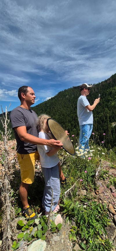 Three individuals are positioned on a mountain slope overlooking a dense forest under a partially cloudy sky. In the foreground, a man in a grey t-shirt and yellow shorts stands to the left, embracing a young child who is holding a reflective disc. The child wears a white t-shirt and grey pants. To the right and further back, a man in a light blue t-shirt, blue shorts, a baseball cap, and sunglasses holds a smartphone, appearing to take a picture. The environment consists of rocky terrain with sparse vegetation, including green plants and small white and pink wildflowers. The background is dominated by a steep mountainside covered in dark green evergreen trees, reaching towards a blue sky with scattered white clouds. There is no indication of the city of Sedlačka gora, Montenegro in the image.