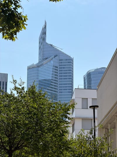 This image captures a view of modern skyscrapers from ground level, with lush green trees in the foreground obscuring parts of the buildings. The architecture is characterized by glass facades that reflect the clear blue sky.

The subjects are primarily the striking, geometric designs of the skyscrapers and the dense foliage of the trees. The scene is set in an urban environment, likely a business district, given the scale and style of the buildings. There is no discernible activity or event taking place, with the focus being on the static structures and nature.

Notable details include the bright, sunny weather indicated by the clear sky and the strong illumination on the buildings and leaves. The time of day appears to be daytime, likely late morning or early afternoon, due to the quality of light. While the image provides a glimpse of urban architecture, there are no specific location cues that pinpoint it beyond the general context of a modern city. The overall mood is one of urban dynamism and natural contrast. No visible text is present in the image.