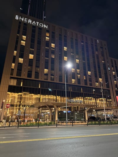 This media file captures a nighttime exterior view of the Sheraton Hotel in Astana, Kazakhstan. The imposing, modern hotel building dominates the frame, with its facade featuring numerous windows reflecting the warm, golden interior lights. The building is tall and appears to be constructed with glass and light-colored materials.

Prominently displayed on the side of the hotel is the "SHERATON" logo in large, illuminated white letters. Adjacent to the hotel, a street or roadway is visible, marked with yellow lines. A black metal fence separates the hotel entrance area from the street, with a grassy strip and some decorative elements behind the fence.

The scene is illuminated by the hotel's internal lighting and streetlights, creating a well-lit but dark nighttime atmosphere. The sky is dark, suggesting it is late at night or early morning. There are no visible people clearly identifiable, but reflections in the windows hint at some activity inside. A red and white circular sign, likely a traffic or informational sign, is visible near the entrance. The overall impression is one of a well-lit, upscale urban hotel at night.
