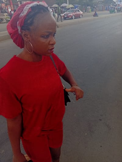A woman in a red shirt and shorts stands on an asphalt road. She wears a red head covering with white lace. Gold hoop earrings are visible. She has a black purse slung over her shoulder. The background shows a street scene with pedestrians, parked cars, and buildings under an overcast sky.