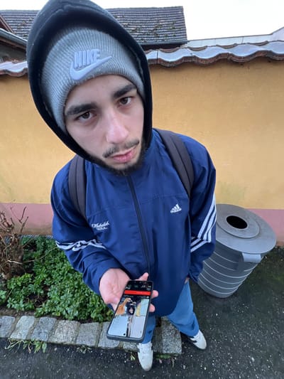 A young adult male stands outdoors in Meistratzheim, France, looking directly at the camera from a high-angle perspective. He has a dark beard and mustache and is wearing a dark navy blue hooded jacket with white stripes (Adidas logo visible), a gray knitted beanie with a white "NIKE" logo, blue jeans, and white sneakers with black stripes. A dark backpack is visible on his shoulders. In his right hand, held at waist level, he displays a black smartphone horizontally, which shows an image of two individuals and a user interface with red and white elements.
The immediate ground is a wet, dark paved surface, bordered on the left by cobblestones and green leafy bushes, along with a small, bare-branched plant. Behind the subject, a light yellow/orange stucco wall extends across the frame. To the subject's right, a gray, ribbed cylindrical public trash receptacle with a black opening is present. Above the wall, a tiled roofline with a gutter system is visible, and the upper background consists of an overcast, light gray sky.