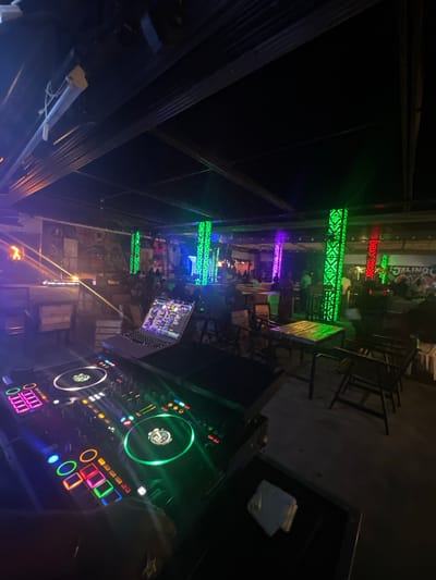 The image depicts the interior of a dimly lit establishment, likely a club or bar, in Howayi, Nigeria. In the foreground, a DJ setup is visible, featuring a laptop with a colorful display, a DJ mixer with illuminated buttons and turntables, and what appears to be a hand with a ring interacting with the equipment. The background reveals a seating area with tables and chairs, and patrons are dimly discernible. The environment is decorated with vibrant, colorful LED lighting, including green, purple, and red accents on pillars and along the ceiling. Murals or artwork adorn the walls. The overall atmosphere suggests a nightlife setting with entertainment being provided.
