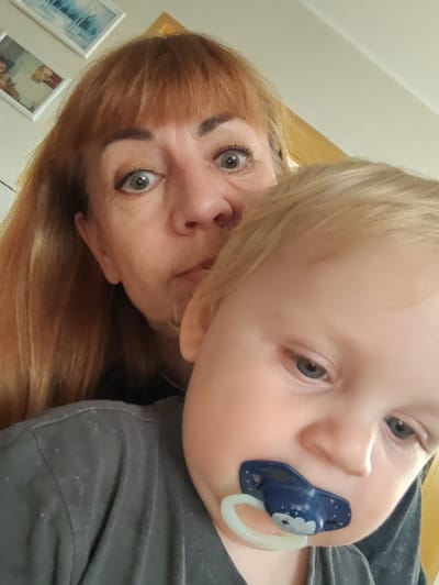 A woman with auburn hair and bangs is taking a selfie with a baby. The woman's eyes are wide, and she is looking directly at the camera. The baby has blond hair and a blue pacifier in its mouth. The background is a wall with two framed pictures hanging on it. The room appears to be indoors. The image was taken in the city of Gvardeysk, Russia.