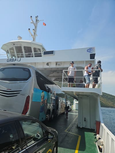 In Lepetane, Montenegro, a ferry with the name "GRBALJ" is seen docked or in the process of boarding. The ferry's deck is filled with vehicles, including a silver bus adorned with an image of people wearing scuba gear and a dark-colored car in the foreground. Several people are visible on an upper deck, including a young person wearing a hat and white t-shirt, and three men standing and looking outwards. One man wears a white tank top and denim shorts, another a black and white patterned shirt, and a third is partially visible. A "FREE Wi-Fi" sign is mounted on the side of the ferry. The sky is clear and blue, and the water is calm, with green, tree-covered hills visible in the background. The scene depicts a typical ferry crossing operation in a coastal town.