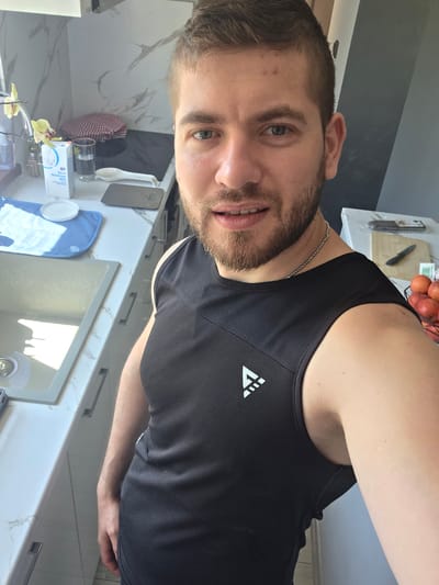 A selfie of a Caucasian male with brown hair, stubble beard, and light skin. He is wearing a black sleeveless athletic shirt with a white logo on the upper left side. He appears to be standing in a kitchen with a marble countertop, sink, kitchenware, and appliances visible in the background. A bowl of red fruits is also visible at the bottom of the frame. There is no information to connect the location to Yagodovo, Bulgaria.