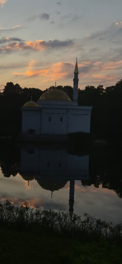 The image depicts the White Mosque (Ak-Mechet) in the city of Pushkin, Russia, at dusk. The mosque, a white structure with golden domes and a minaret, is situated on the edge of a body of water. The setting sun casts a warm glow on the clouds in the sky, creating a colorful and dramatic backdrop. The mosque is reflected in the still water, creating a symmetrical and serene scene. Trees surround the mosque and line the edges of the water, their dark silhouettes contrasting with the lighter colors of the sky and the mosque. The foreground is occupied by dark, silhouetted vegetation. The overall atmosphere is peaceful and tranquil.