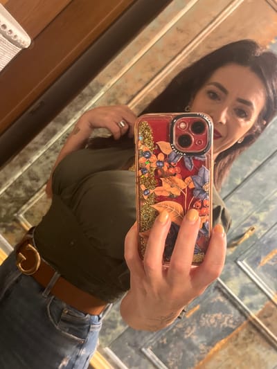 A woman with dark hair is taking a selfie in a mirror. She is wearing a dark green top and blue jeans with a brown belt. Her phone is decorated with a floral design and glitter. The background appears to be a bathroom with framed mirrors. The location is not possible to determine without further information.