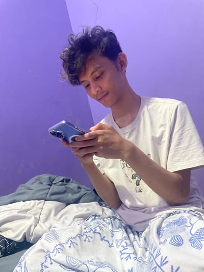 A young person with curly dark hair is sitting on a bed, looking down and using a smartphone with a blue case. The person is wearing a white t-shirt with a graphic print. The bed is covered with various blankets and bedding, some of which have blue and white patterns. The wall behind the person is painted purple. The lighting suggests an indoor setting, possibly a bedroom, with a soft, ambient glow, likely from a lamp or LED lighting. The scene does not contain any explicit indicators of location within North Jakarta, Indonesia, but depicts a common domestic environment.