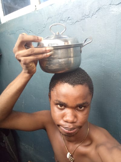 A man is balancing a metal pot on his head. He has short, dark hair and a gold necklace. He is shirtless. The background is a blue wall. The location is likely Owerri, Nigeria.