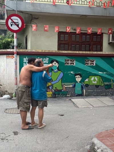 Two men embrace in front of a mural on a street in Hanoi, Vietnam. The mural depicts a green garbage truck and two figures. One figure, wearing a blue shirt and mask, is handing something to another figure wearing a green shirt. The mural is painted on a green wall with white and yellow stripes.
The two embracing men are dressed casually. The man in front, facing away from the camera, wears a blue t-shirt and khaki shorts. He has glasses. The man behind him, whose back is to the camera, is shirtless and also wears khaki shorts. They are standing on a gray asphalt street.
To the left of the mural, there is a building with wooden-shuttered windows and small red flags strung along the facade. A red and white circular sign with a diagonal line through it, indicating "no entry" or a prohibition, is visible in the foreground. On the right side of the image, part of a sidewalk with red brick accents and some foliage can be seen.
The scene appears to be set during the daytime, with overcast or diffused lighting. The overall atmosphere suggests a moment of affection or comfort shared between the two men in a public space decorated with cultural and political symbols. Visible text on the mural is difficult to decipher clearly, but it appears to be in Vietnamese. There is also text on a signpost to the left of the men, reading "MAM NON IQ" and some Vietnamese characters.