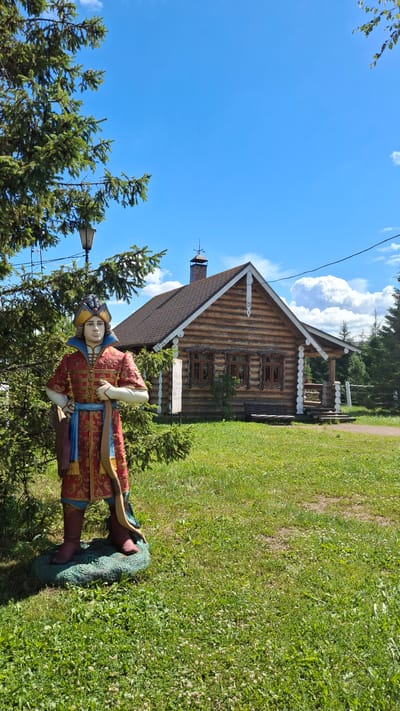 This media file depicts a scene on a bright, sunny day, likely during daylight hours given the clear blue sky and strong shadows. The setting is an outdoor area with abundant green grass and mature trees, suggesting a park or a historical village. In the foreground, a statue of a person dressed in traditional, ornate clothing stands on a circular, teal-colored base. The figure is wearing a red garment with gold and blue embellishments, a blue sash, and a distinctive headdress.

Behind the statue, a traditional wooden house, possibly a log cabin, is visible. The house features intricate wood carvings around the windows and a prominent chimney. The roof is shingled, and there's a porch area with white railings. A lamppost is also present in the scene, situated to the left of the statue and in front of the house.

The overall impression is of a cultural or historical site, perhaps an open-air museum, given the presence of the statue and the rustic architecture. The weather appears pleasant, with a few white clouds scattered across the vibrant blue sky. The location is identified as Peterhof, Russia, which further supports the interpretation of a historical or cultural attraction in that area. There is no discernible text visible in the image.