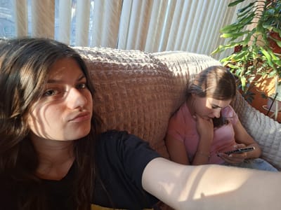 Two young women are on a sofa. The woman in the foreground has long brown hair, a black shirt, and is taking a selfie. Sunlight from the window blinds falls on her face. Behind her to the right, the other woman, also with brown hair, is wearing a pink shirt and light blue jeans. She is looking down at a phone she holds in her hands. The sofa is a textured, light brown color. Behind them are vertical window blinds and a large leafy plant in a pot. The location is Panagyurishte, Bulgaria.