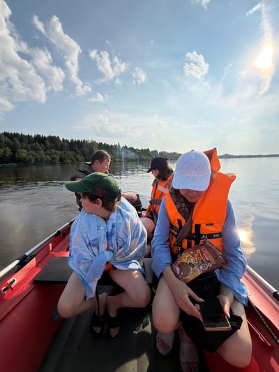 Four people are in a red motorboat on a body of water. The boat is moving, as indicated by the wake behind it. The sky is partly cloudy with the sun visible. On the left side of the boat, a person wearing a green baseball cap and a blue and white patterned garment is looking to their left. Behind them, another person in a green vest is looking forward. On the right side of the boat, a person wearing a white cap and an orange life vest is looking down at a phone and a snack bag. Next to them, another person in an orange life vest is also looking forward. The boat is passing a densely wooded shoreline. The scene appears to be in a rural area.
