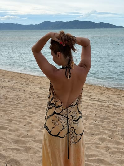 A woman with auburn hair tied up stands on a sandy beach facing away from the camera, looking out at the ocean. She is wearing a flowing, sleeveless, light-colored dress with a black tree-like pattern. The dress has a deep V-back and is tied at the neck with black straps that have a decorative knot. Her arms are raised, and she is adjusting her hair with her hands. The ocean is a muted blue with small waves, and in the distance, a line of dark mountains is visible under a cloudy sky. The sand on the beach is a light brown, with subtle ripples from the wind or water. The scene appears to be a serene coastal setting in the Ko Samui District of Thailand.