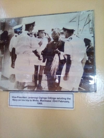 The image is a photograph of an older, framed black and white picture, dated February 23, 1965. The older picture depicts Vice-President Jaramogi Oginga Odinga interacting with Navy personnel during his trip to Mvita, Mombasa, Kenya. The context of this description includes South Sakwa ward, Kenya.
In the center of the depicted scene, Jaramogi Oginga Odinga, an African man, wears a dark, possibly traditional, long-sleeved garment with buttons down the front, and a dark cap. He holds a long, light-colored, possibly feathered, object, resembling a fly-whisk, in his right hand. He faces towards the right of the frame, engaging with uniformed naval officers. Surrounding Odinga are at least four male Navy personnel in crisp white short-sleeved uniforms with epaulets, dark ties, white shorts, dark socks, and white peaked caps. Two officers are prominent in the foreground, facing Odinga; the one on the left has his left hand extended towards Odinga, possibly in a greeting or handshake. The scene is outdoors on what appears to be a paved area, possibly a dock or ceremonial ground, with indistinct structures and what might be ship rigging or poles visible in the distant background under daylight conditions.