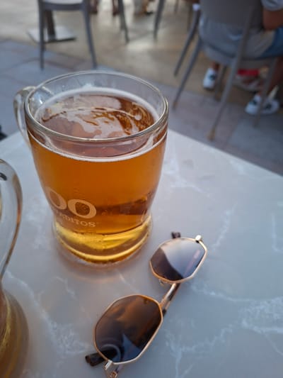The image shows a close-up of a glass of amber-colored beer with foam on top, resting on a marble-patterned table. A pair of gold-rimmed sunglasses with dark lenses is placed next to the glass. In the background, slightly out of focus, are chairs and table bases, suggesting an outdoor seating area of a cafe or restaurant. The lighting indicates it is daytime. Although the specific location within Torrevieja, Spain is not discernible from the image alone, the setting appears to be a typical urban or coastal establishment. There are no people clearly visible or interacting with the objects.