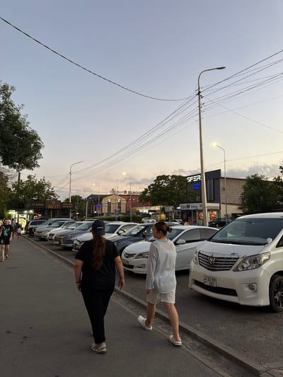 The image depicts a street scene in Shymkent, Kazakhstan, likely during dusk given the soft, diffused light and visible streetlights. The perspective is from ground level, looking up and across the street, with the image rotated 90 degrees counter-clockwise.

Two women are the primary human subjects. One, wearing a light blue shirt and white shorts, is walking away from the viewer and toward the parked cars. The second woman, dressed in a black t-shirt and black leggings, is also walking away from the viewer, slightly ahead of the first woman. She is wearing a dark baseball cap.

Several white and silver cars are parked along the side of the street. One white car in the foreground has a license plate visible that reads "02 KP 087" and appears to be from Kazakhstan. A building with illuminated signs stands behind the parked cars. Some of the visible text on the signs includes "SFER", "БИЛЬЯРД", "КЛУБЫ", "ЕТ ДҮКЕНІ", "МЯСНОЙ МАГАЗИН", and "Vipoj 1 Центр".

Utility poles with numerous overhead wires are prominent in the scene, stretching across the sky, which is a pale, overcast grey with hints of yellow or orange near the horizon. Trees with green foliage are also visible, adding to the urban but naturalistic setting. The overall atmosphere appears calm and ordinary, suggesting a typical evening on a city street.

The image is rich with details of urban life, including transportation, commercial establishments, and infrastructure, set against a soft twilight sky.