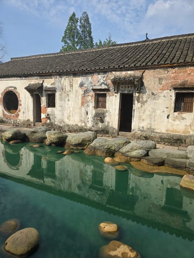 This image shows a weathered, traditional Chinese building with a tiled roof, situated beside a body of tranquil, turquoise water. The building's facade is a mix of crumbling plaster and exposed red brick, with several windows, some barred, and a prominent arched doorway. Large, moss-covered rocks line the edge of the water, some submerged, creating a natural border. The clear water reflects the building and the sky above, along with scattered smaller rocks. In the background, two tall evergreen trees stand against a blue sky dotted with white clouds. The architecture and natural setting suggest a historical or cultural site, possibly in a rural or heritage area within the Xiaoshan District of China. No people are visible in the scene.