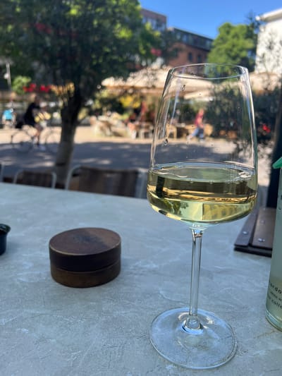 A wine glass filled with white wine sits on a table outside, with a blurry background of trees, buildings, and people. The scene suggests a casual outdoor setting, possibly a cafe or restaurant, on a sunny day. The light creates reflections and highlights on the glass, showing some condensation. The wine is a pale yellow color. To the right of the glass is a small, dark brown, circular object, possibly a lid or coaster. In the upper background, a partial view of a bottle with text is visible. The text reads "chardonnay white wi...e collec...". The overall impression is relaxed and summery. The image is oriented vertically, and the subjects are viewed from above. The lighting indicates it is daytime, likely late morning or early afternoon given the shadows and brightness.