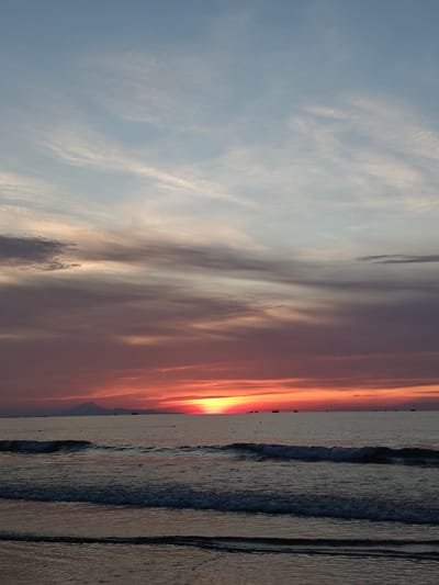 This is a photograph of a sunset over the ocean in Curehtunong, Indonesia. The sky is filled with dramatic clouds in shades of orange, pink, and purple, transitioning to a pale blue at the top. The sun is setting on the horizon, casting a warm glow across the water. Small waves are rolling onto the shore, creating a textured pattern on the wet sand. Several distant boats are visible on the calm sea. In the far distance, a silhouetted mountain range is visible on the left side of the frame. The overall scene evokes a sense of tranquility and natural beauty.