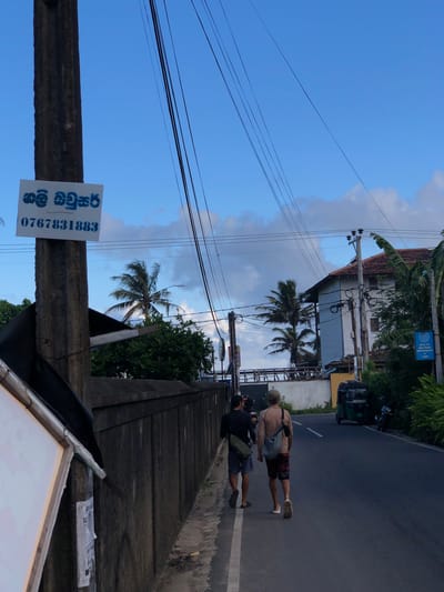 The image captures a street scene in Ahangama, Sri Lanka, likely during the late afternoon or early evening given the soft, diffused light. The sky is a clear blue with scattered clouds, suggesting fair weather. The setting is a road with power lines crisscrossing overhead, indicative of a well-traveled but perhaps less developed area.
In the foreground, a concrete pillar supports a sign with text in what appears to be Sinhala script, along with a phone number: "0767831883". The script is blue and stenciled.
Further down the road, two people are walking away from the camera. One individual, wearing a light-colored shirt and shorts, carries a bag slung over their shoulder. The other person, in darker attire, also has a bag. Their postures suggest they are engaged in conversation or simply walking together.
On the right side of the road, a building with a tiled roof and lush green foliage is visible. A small, three-wheeled vehicle, commonly known as a tuk-tuk, is parked nearby. The overall impression is that of a typical day in a coastal town in Sri Lanka, with everyday activities unfolding along the roadside. The scene is calm and unhurried, with no overt signs of distress or urgency.